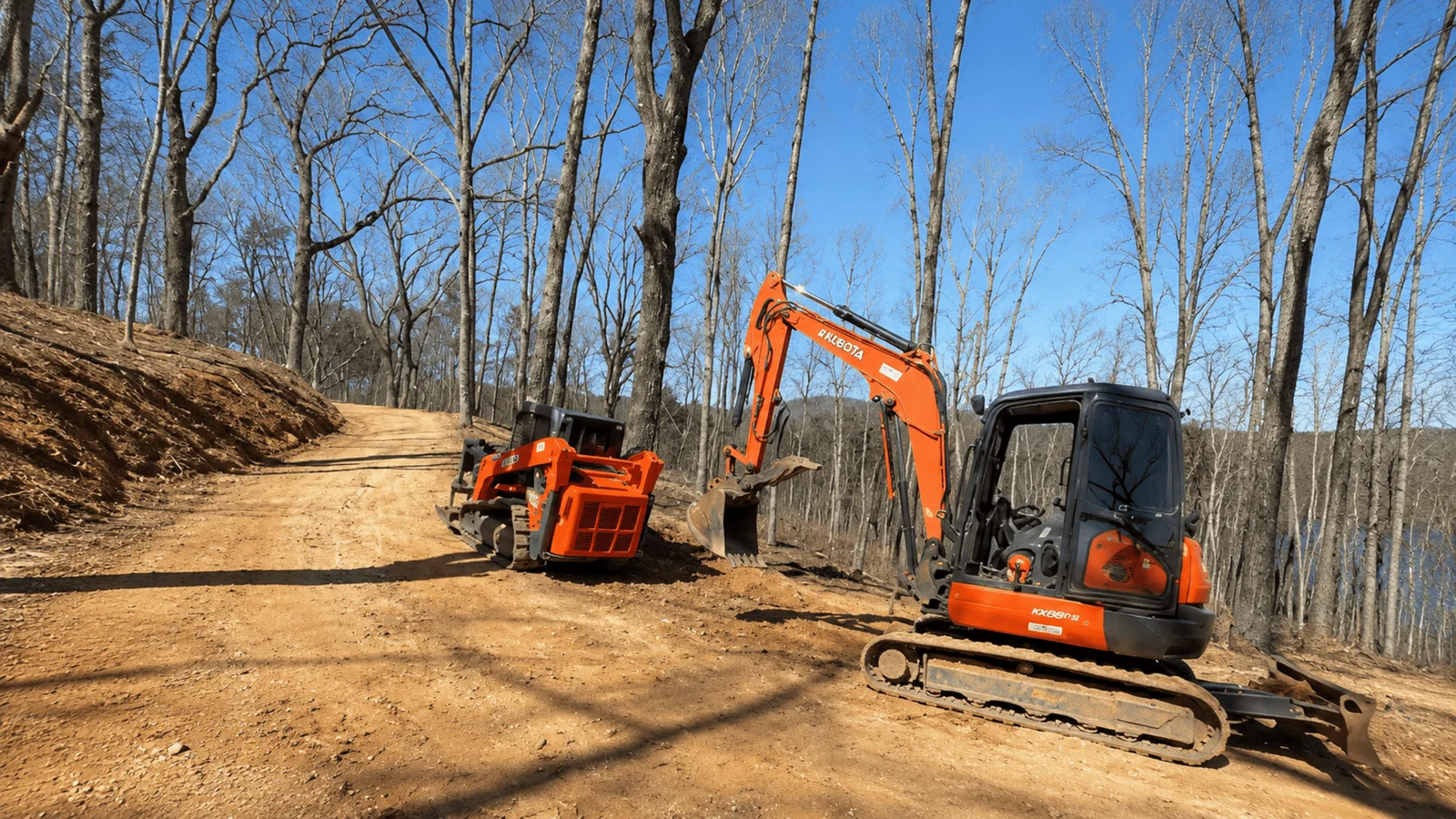 Compact excavator and skid steer with bucket on cleared dirt site surrounded by bare trees
