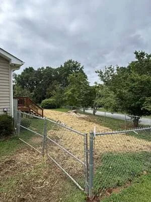 Cleared residential lot with chain-link fence, graded soil, and mature trees retained