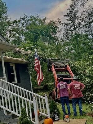 Bucket truck with extended boom removing tree limbs near residential house with white railings