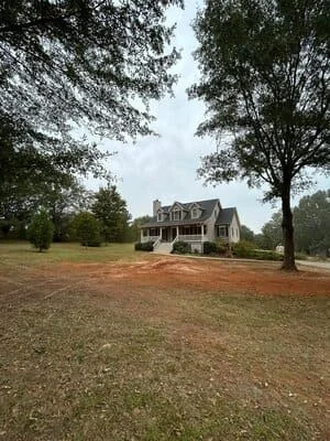 Cleared residential lot with removed trees, mature oaks remaining, graded soil around two-story home